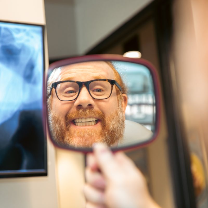 Patient with glasses smiling while checking his teeth in a handheld mirror with a dental X-ray in the background.