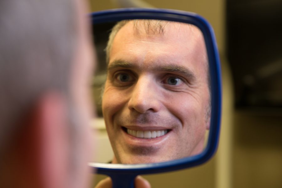 Man smiling and looking at his reflection in a handheld mirror.